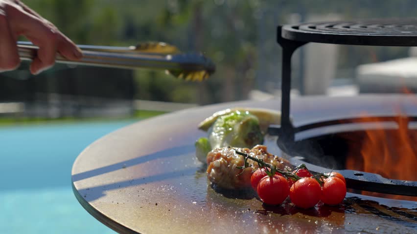 close-up of a brazier with a hand holding tongs and turning food on the hotplate, flame in the centre of the brazier and blurred swimming pool in the background