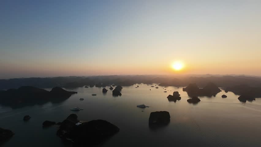 Cinematic aerial drone footage featuring a smooth panoramic pan across Ha Long Bay, Vietnam, captured during a stunning sunset. The warm golden-hour light highlights towering limestone karst formations and tranquil emerald waters, creating a breathtaking seascape within this UNESCO World Heritage Site. Perfect for travel films, tourism promotions, documentaries, and scenic landscape projects.