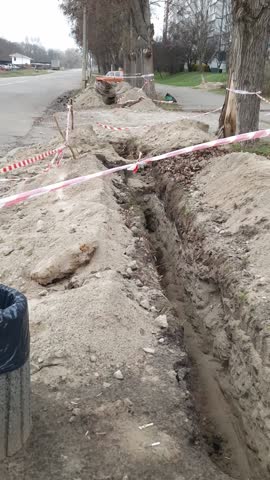 Deep earthen trench dug for utility pipes or cables in an urban park area. Red and white warning tape marks the hazardous construction zone.
