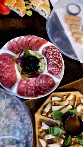 Assortment of appetizers including a salami platter, sushi rolls, and a cheese board are arranged on a wooden table. Close-up overhead view reveals the abundant and appetizing food display.