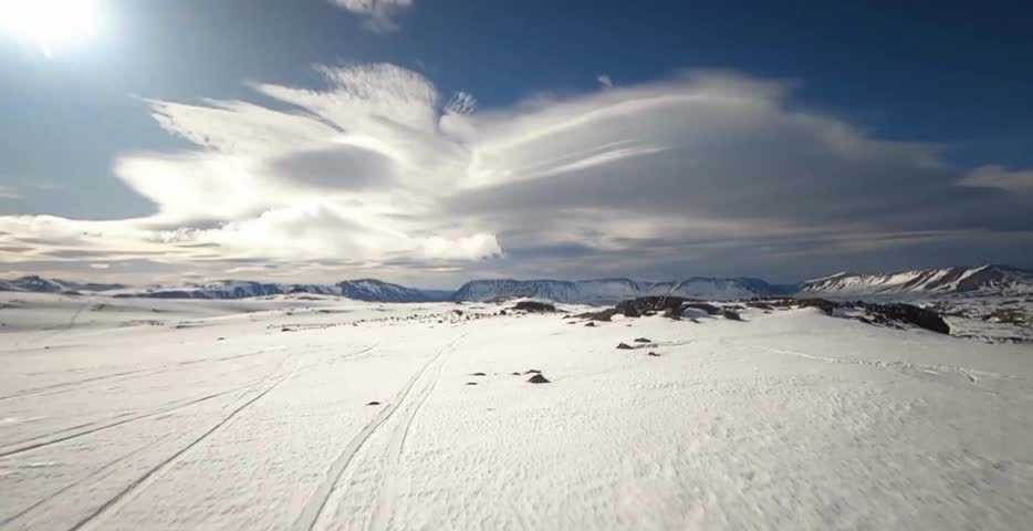 Aerial view of a snow-covered alpine hillside with scattered pine trees and a deep blue mountain lake below. Bright winter light highlights pristine nature and dramatic elevation.
