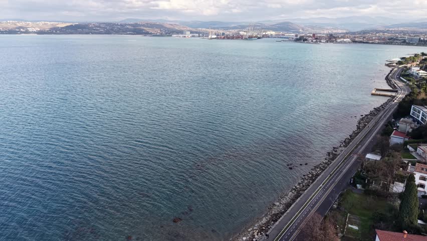 Cinematic drone panorama over the Bay of Koper on the Adriatic Sea. Blue-green water with gentle ripples, rocky shoreline and the coastal road in the foreground, with the Port of Koper cranes and cityscape in the distance