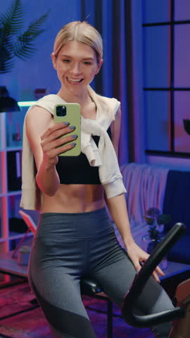 Portrait of charming cheerful athletic fit slender 25-aged blonde in training clothes which photographing herself on smartphone while turning the pedals on bike trainer in cozy night lighted room