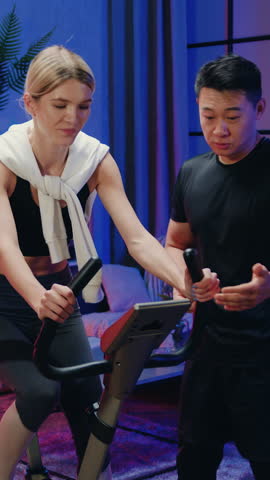 Joint home workout concept where attractive fit sportive slender young woman in sportswear riding on bike trainer while her likable strong positive asian boyfriend pressing the buttons on control