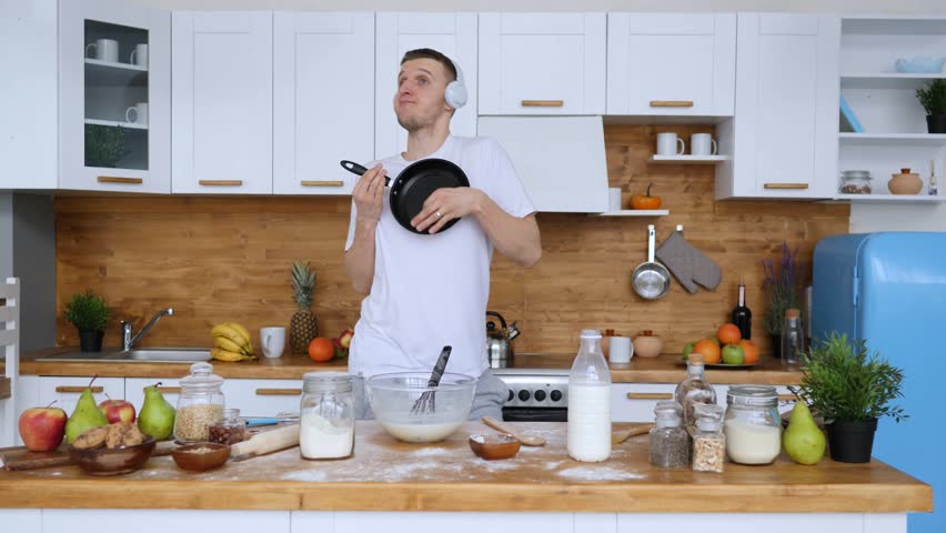 Cheerful young man wearing headphones listening to music and dancing while preparing breakfast, playfully pretending to play a guitar with a frying pan in a modern, spacious kitchen at home