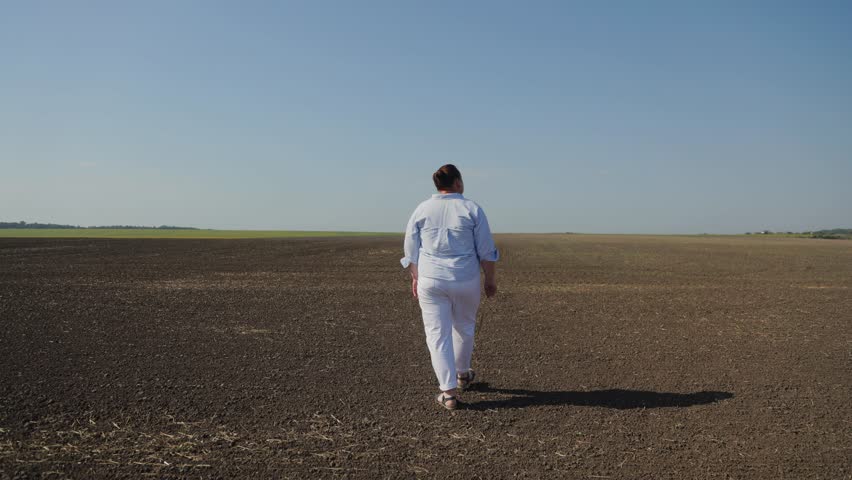 Solitary woman walking on a large, empty plowed field under a clear blue sky