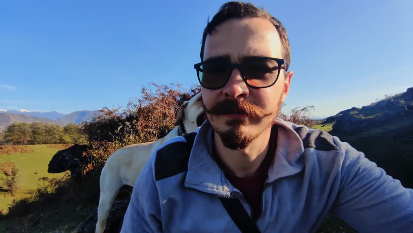 man with dog outdoors hiking through golden meadow at sunset, wearing sunglasses and backpack, labrador leaning close panting with tongue out and licks a persons cheek, candid closeup portrait