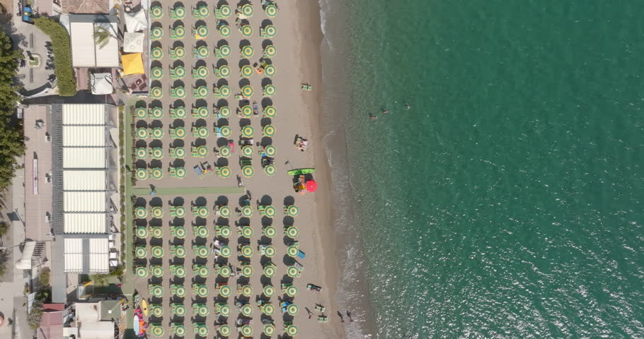 Top-down aerial shot of a beach with neat rows of green umbrellas. A red lifeguard station stands out on the sand next to the clear blue sea where people are swimming. Zenith view of Sicily, Italy.