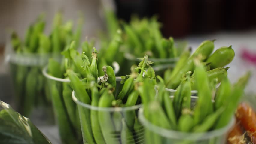 Close-up view of fresh green snap pea pods standing upright in clear plastic cups on a produce stall, highlighting vibrant color, crisp texture, and farm-fresh market abundance.