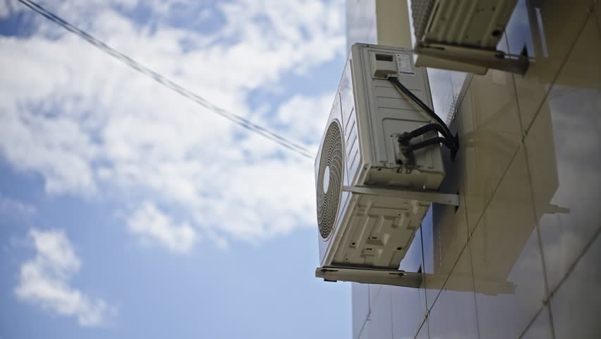 Outdoor wall-mounted HVAC condenser unit on modern building facade under blue sky, showing cooling equipment, brackets, wiring, and ventilation components in low-angle perspective