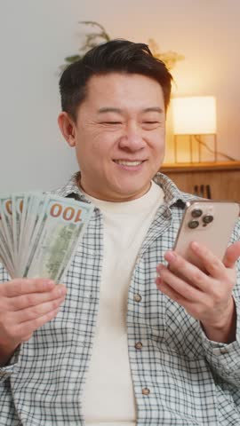 Successful rich man holding smartphone and waving dollar bill money fan sitting on sofa couch at home. Happy Chinese guy male winning online lottery game planning vacation calculating budget concept.