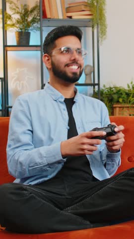 Cheerful Indian young man using joystick controller playing video game fun enjoying sitting on sofa in living room. Arabian guy enjoying success winning online game on couch during weekend at home.