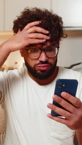 Arabian man at home checks smartphone gets bad news game loss lottery frowns shakes head no sighs. Young guy on sofa reads message about result looks upset frustrated sad reacts to negative alert.