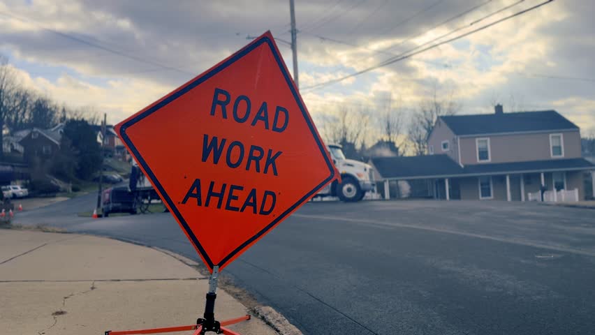 Bright orange road work ahead sign on a tripod beside a paved street, warning drivers of construction and traffic changes ahead. Pittsburgh suburbs. 15012  