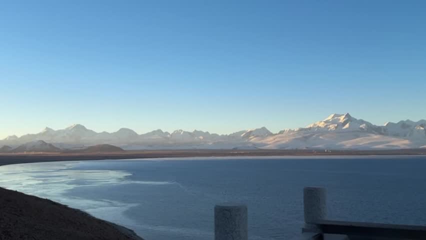 Horizontal scenic footage of a vast high-altitude alpine lake with deep blue water. A majestic snow-capped mountain range stands in the background under a clear sky. The water surface ripples gently in the cold wind. Landscape likely in Tibet or Qinghai, China (Namtso or Yamdrok Lake). 4K video.