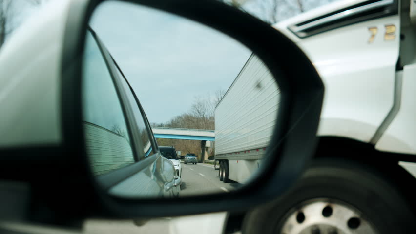 POV view. Reflection of white truck in the side mirror of driving car on the road on spring day. View from front passenger seat. Clouds are in the mirror.