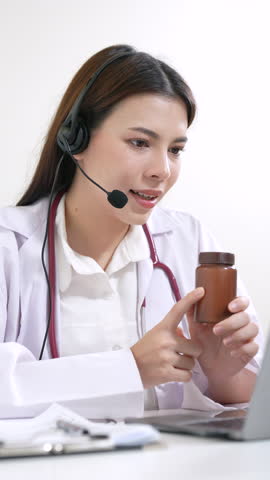 Asian woman doctor in headset examining medicine bottle during telemedicine session, concentrating on dosage instruction, online healthcare, digital consultation, and professional medical service