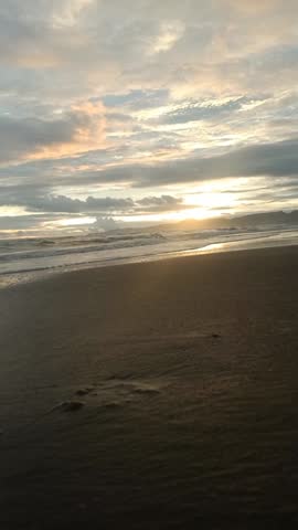 A serene sunset over a sandy beach with gentle waves washing ashore. The golden hour light reflects off the clouds and wet sand, creating a peaceful and moody atmosphere