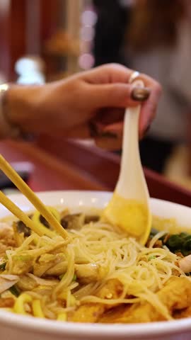 A close-up vertical view of a person lifting thin noodles from a savory broth.