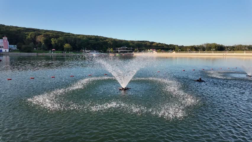 Fountains spray water in park lake at sunset with trees in background and calm water surface