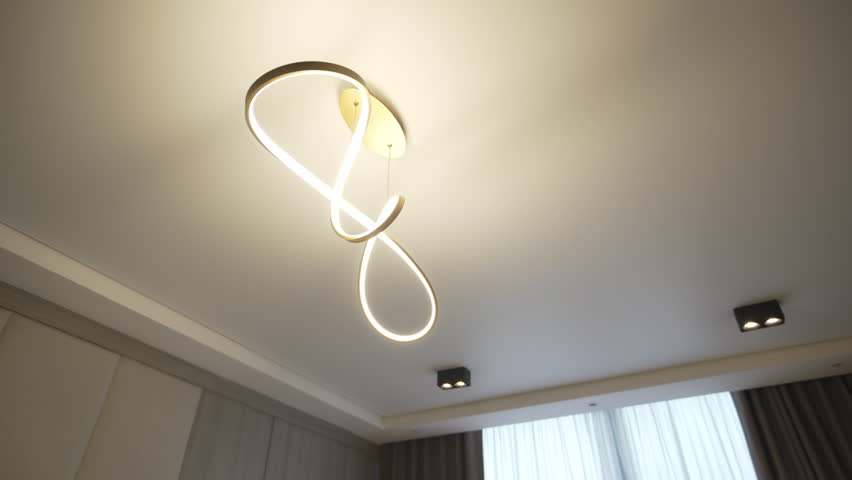 Ribbon shaped ceiling chandelier forms flowing luminous sculpture against light ceiling surface. Lighting composition creates soft glow