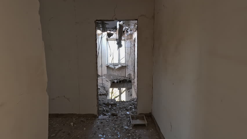 Extensive structural damage to a residential building featuring holes in the roof and ceilings, blown-out doors, and shrapnel marks on walls after an airstrike