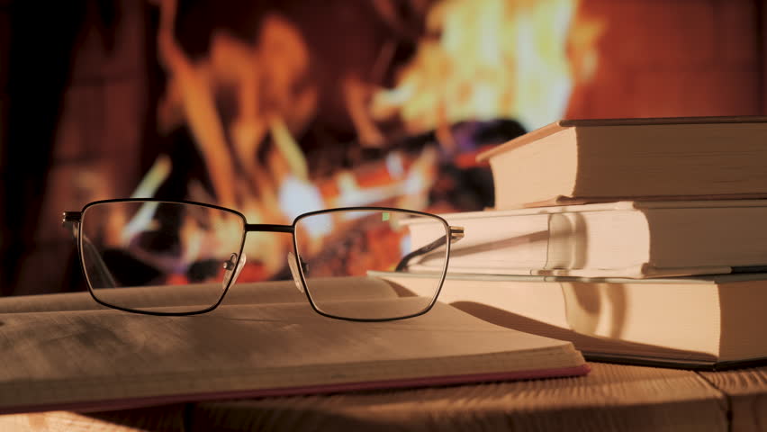 Eyeglasses placed on book stack beside burning fireplace, reading optics resting near hardcover volumes with warm indoor atmosphere