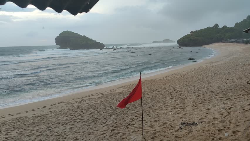​A rainy day at a tropical beach with grey skies, choppy waves, and a red warning flag standing on the sand.