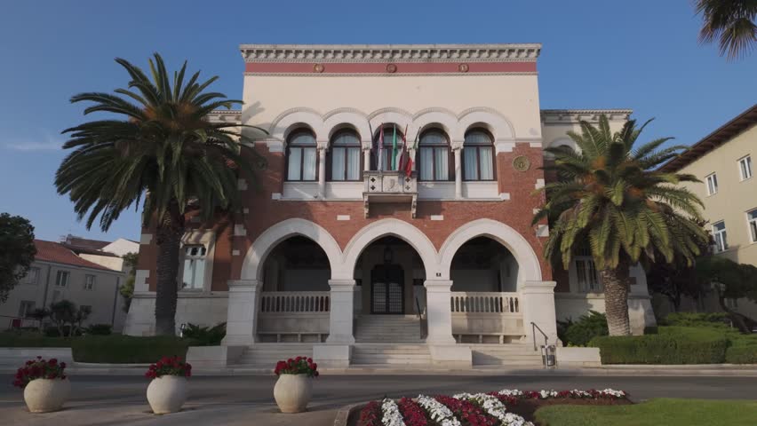 The beautiful Town Hall of Porec, Croatia on a sunny summer afternoon. Built in 1910, designed by an Italian architect. Romantic street scenery with huge old palm trees, and red and white flowers.
