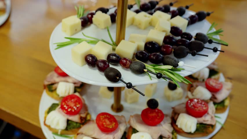 Multilevel serving tray displaying gourmet appetizerscheese cubes, olives, grapes, canapes with pesto, ham, cherry tomatoes, complementing upscale gathering ambiance