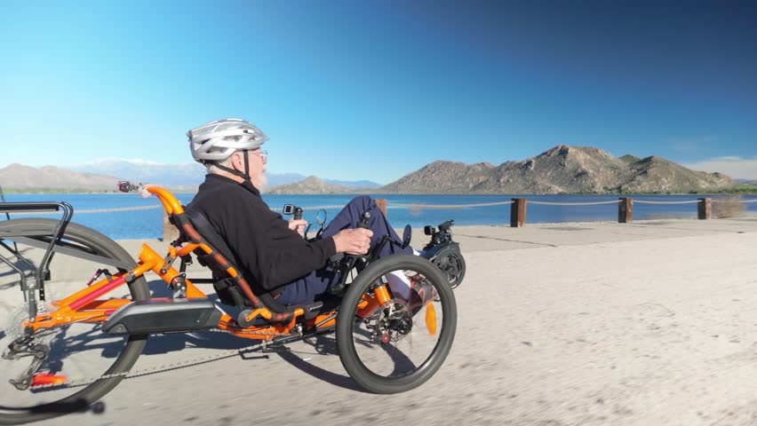 Senior man enjoys biking on a recumbent electric trike along a lakeside path with mountains in view under a clear sky