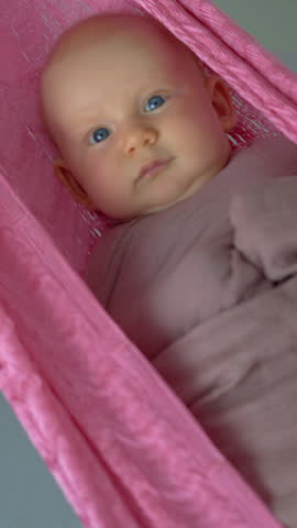 Loving mother gently swings her swaddled two-month-old baby in a hammock to help him fall asleep, creating a peaceful and nurturing family bedtime moment.