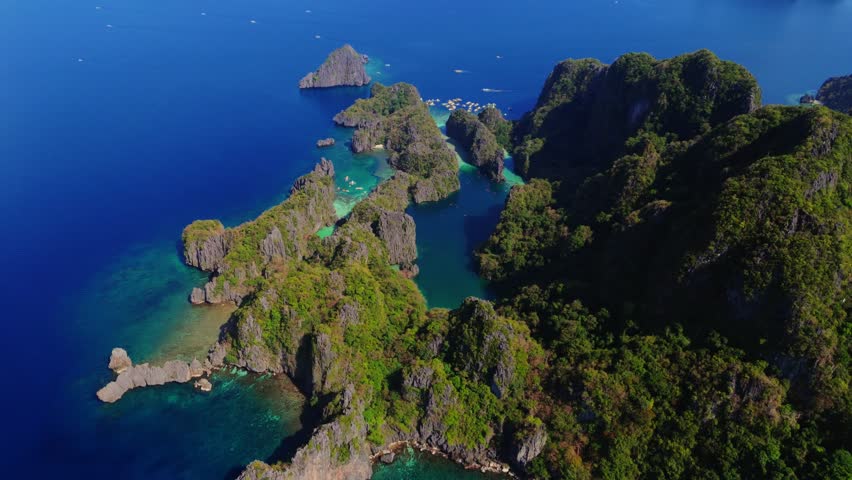 Aerial view of jagged islands with lush green vegetation, surrounded by turquoise and deep blue waters dotted with boats, El Nido, Philippines.