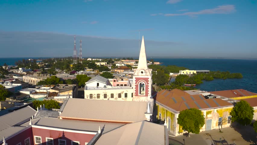 Aerial view of a church with a tall spire and colorful buildings by the ocean, reflecting the clear blue sky, Nampula, Nampula Province, Mozambique.