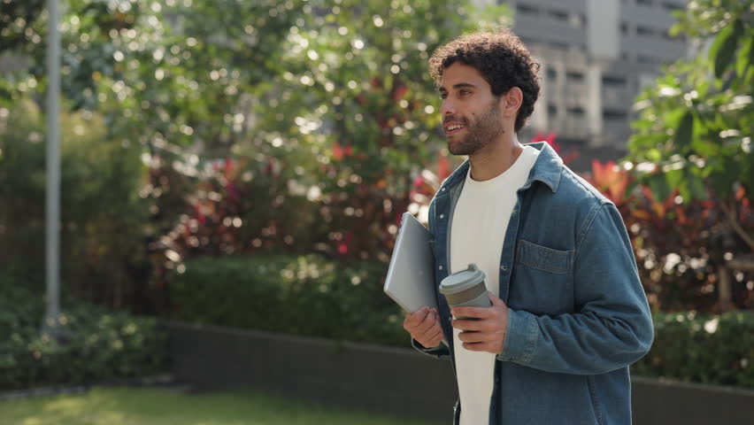 Portrait of happy Hispanic man with digital tablet drinks coffee in green city park. Latin male student holds mobile device as walks along spring downtown street slow motion