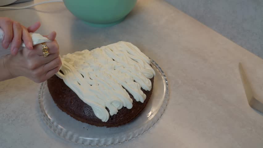 Woman decorating a chocolate sponge cake by piping fresh cream frosting from a pastry bag on a glass plate in the kitchen, preparing homemade dessert.
