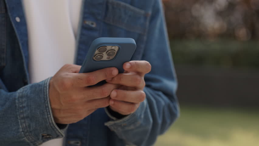 Closeup view of man hands type message on smartphone in spring city park. Young male in denim casual clothes writes post in social media by mobile phone as rests in summer city