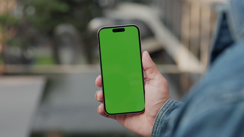 Closeup view of man hand holds smartphone with green chromakey screen for advertisement in modern city. Male shows cellphone with blank mockup display as stands in downtown