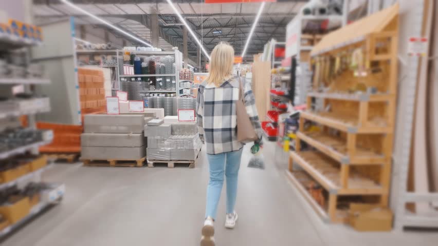 Blonde woman wearing a plaid shirt walking down an aisle in a large hardware store, looking for building materials and supplies for home improvement, renovation, or do-it-yourself projects