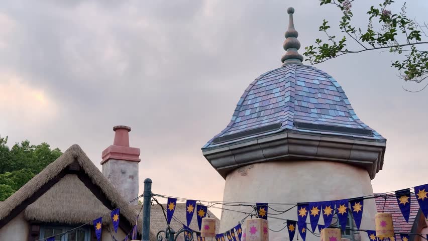 A whimsical tower roof and pastel shingles rise above strings of lantern banners. Dusk sky and storybook village details set a fairytale mood in Orlando, Florida.