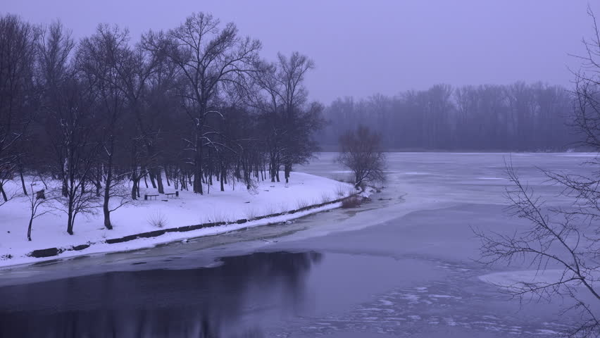 Misty winter landscape showing a frozen riverbank with bare trees reflected in the dark water. The snowy shoreline and tranquil atmosphere create a serene. Cold. And picturesque scene in a city park
