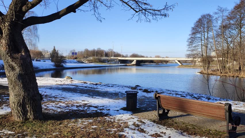 There is a wooden bench on a metal base and an urn on a tiled path near a tree. There is a concrete road bridge over the river and cars are driving. Sunny winter weather and remnants of snow