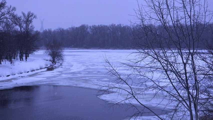Misty winter landscape showing a frozen riverbank with bare trees reflected in the dark water. The snowy shoreline and tranquil atmosphere create a serene. Cold. And picturesque scene in a city park