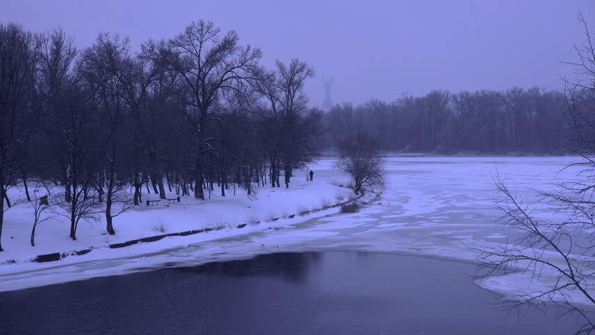 Misty winter landscape showing a frozen riverbank with bare trees reflected in the dark water. The snowy shoreline and tranquil atmosphere create a serene. Cold. And picturesque scene in a city park