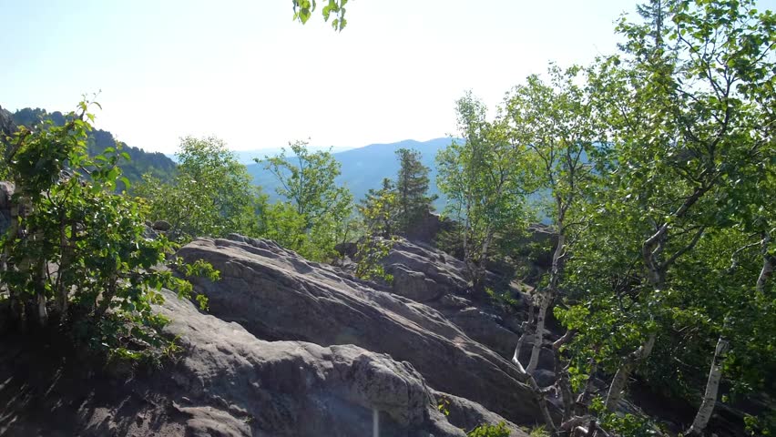 Footage showing a scenic view of rocky mountain slopes covered with green trees and vegetation, overlooking a mountain range in the distance under a clear blue sky.