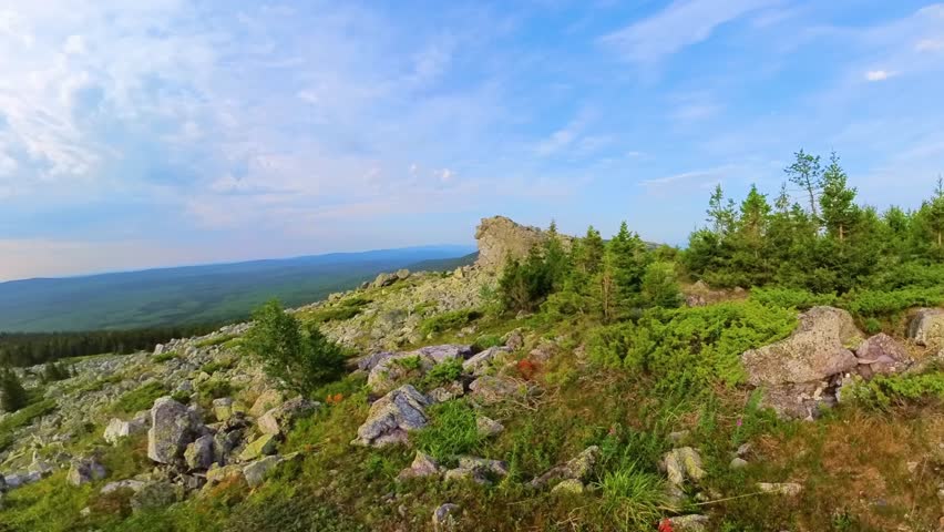 Panoramic view from the summit of a mountain ridge in the South Ural region of Russia. The landscape features large rock formations, green vegetation including small trees and moss, under a partly clo