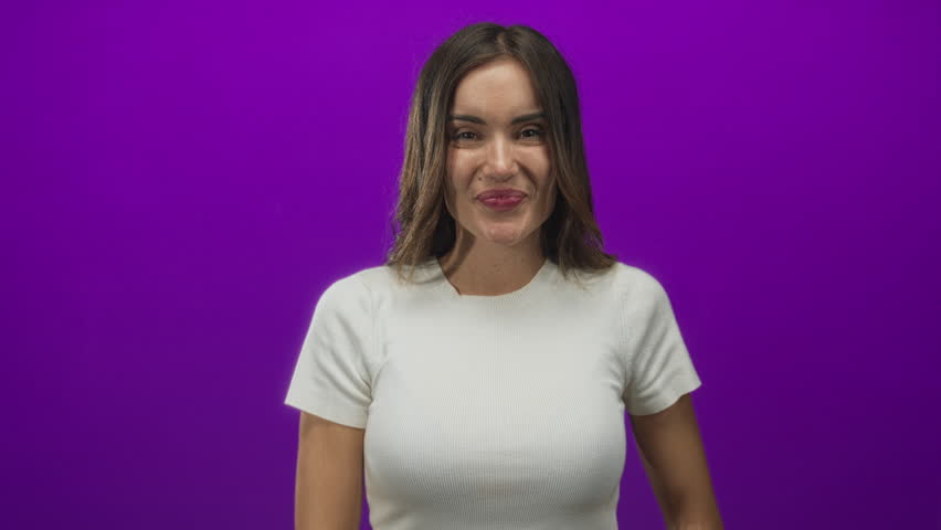 Woman smiling, points finger to temple with gentle head tilt in purple studio wearing white shirt; playful confidence.