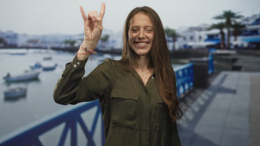 Woman smiles while making rock horns with her hand on a seaside street promenade by a blue railing and moored boats; joy travel freedom.