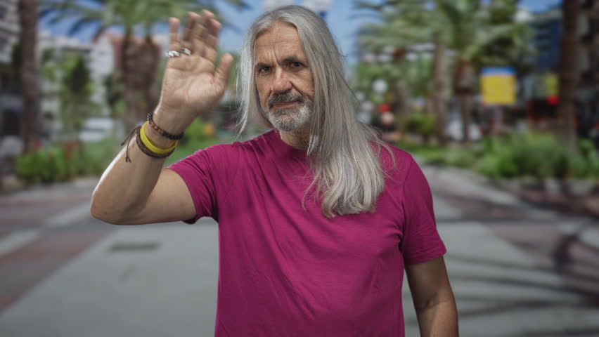 Man with long gray hair waving hand and shielding eyes on a sunny palm lined street promenade wearing bracelets and magenta tshirt; welcoming.