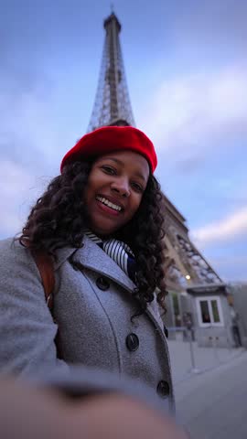 Vertical. African millennial smiles at the camera while taking a selfie beside the Eiffel Tower in Paris at blue hour, wearing a red beret and coat, capturing urban travel vibes with direct gaze.
